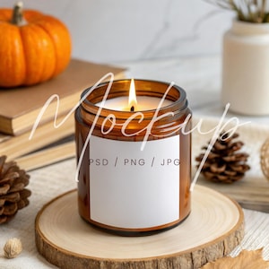May include: A lit candle in a brown glass jar with a white label, set on a wooden slice. An orange pumpkin, books, and pine cones are in the background. The candle flame is bright, and the scene evokes a cozy autumn theme.