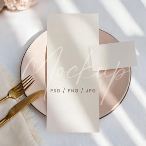 Peut inclure: Mise en scène avec une assiette rose pâle, une fourchette et un couteau dorés, et des cartes blanc cassé. Une grande carte est dressée, avec une petite carte à côté. Le mot "Mockup" est superposé. Sur une nappe blanche.