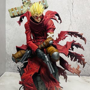May include: A detailed figurine of a character in a red coat, holding two pistols. The figure has spiky blonde hair and red sunglasses. The base is a weathered, textured design with a cross behind the figure.