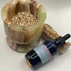 Puede incluir: Un mortero y una maja de &oacute;nix verde, marr&oacute;n y blanco llenos de virutas de madera, junto a una botella de vidrio azul con un cuentagotas. La botella tiene una etiqueta con texto. La maja tambi&eacute;n es de &oacute;nix.