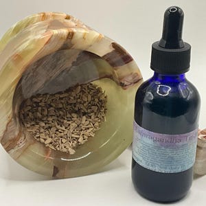 Puede incluir: Una botella de vidrio azul de tintura de Ashwagandha con un cuentagotas negro se encuentra junto a un cuenco de &oacute;nix tallado lleno de virutas de madera. Tambi&eacute;n hay un peque&ntilde;o coraz&oacute;n de &oacute;nix.