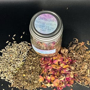 May include: A glass jar of herbal tea blend labeled "Fret Off Tea" with a variety of dried herbs and flowers. The tea is surrounded by piles of lavender, rose petals, and other dried plants. The jar's label includes directions.