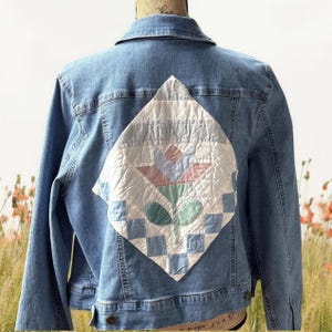 Peut inclure: Une veste en jean bleu clair avec un grand empiècement matelassé en forme de losange dans le dos. L'empiècement présente un fond blanc avec un motif floral central en rose et vert doux, entouré d'une bordure de carrés bleu clair et blancs.