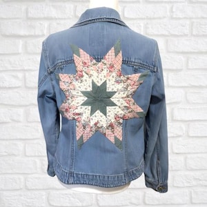 May include: Pale blue denim jacket featuring a large star design on the back. The star is made of fabric with floral and geometric patterns in pink, green, and white. The jacket has a classic collar and button closure.