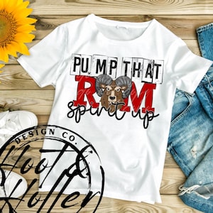 May include: White t-shirt with the text "PUMP THAT RAM spirit up" in black and red. The graphic features a ram's head. The shirt is on a wooden surface with a sunflower and a pair of white sneakers.
