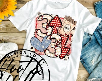 Puede incluir: Camiseta blanca con un diseño rojo, blanco y azul que presenta el número 3, un corazón y una flecha. El diseño también incluye una bolsa de cacahuetes con la palabra "PEANUTS" impresa en ella.