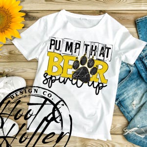 May include: White t-shirt with the text "PUMP THAT BEAR spirit up" in black and yellow. A black paw print is in the center of the design. The shirt is on a wooden surface with a sunflower and a pair of white sneakers.