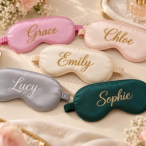 Könnte beinhalten: Fünf Schlafmasken aus Satin in Pink, Hellrosa, Creme, Grau und Dunkelgrün. Jede Maske hat ein elastisches Band und ist mit einem Namen in Schreibschrift bestickt: Grace, Chloe, Emily, Lucy und Sophie. Die Masken haben eine glatte Oberfläche.