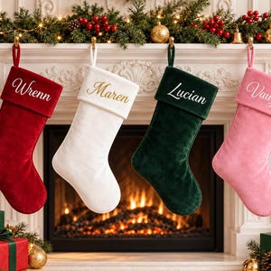 Könnte beinhalten: Vier Weihnachtssocken aus Samt hängen in einer Reihe. Die rote Socke ist mit dem Namen Wrenn bestickt, die weiße mit Maren, die dunkelgrüne mit Lucian und die rosa Socke mit Vaughn. Jede Socke hat einen passenden Umschlag und eine Schlaufe.