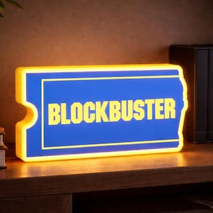 May include: A rectangular Blockbuster sign with a blue background and yellow text. The sign has a yellow border and is lit up. The sign is on a wooden shelf with a plant and books in the background.