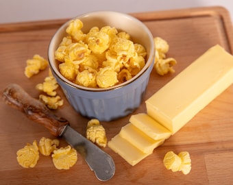 Może przedstawiać: Jasnoniebieska miska wypełniona żółtym popcornem, kostka masła i nóż do masła na drewnianej desce do krojenia. Kilka kawałków popcornu jest rozrzuconych wokół miski i noża.