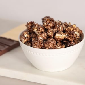 May include: A white bowl filled with chocolate-covered popcorn sits next to a dark chocolate bar. The popcorn is a mix of light and dark brown, and the chocolate bar is partially unwrapped.