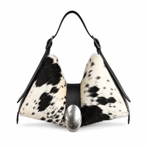 May include: A structured handbag featuring a black and white cowhide pattern on two triangular side panels. A central black leather strap connects the sides, accented by a large, smooth, oval-shaped silver-tone metal buckle. It has a black handle.