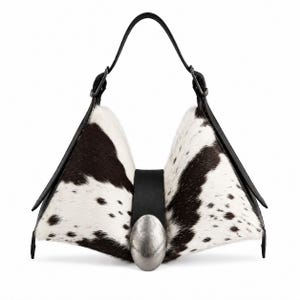 May include: A structured handbag featuring a black and white cowhide pattern with a triangular silhouette. The bag includes a central black leather strap secured by a large, oval-shaped, brushed silver-tone metal accent and a single black handle.