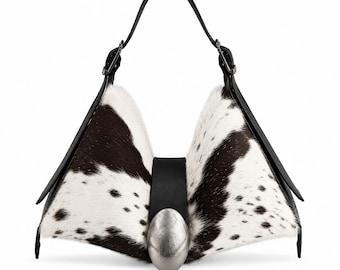 May include: A structured handbag featuring a black and white cowhide pattern with a triangular silhouette. The bag includes a central black leather strap secured by a large, oval-shaped, brushed silver-tone metal accent and a single black handle.