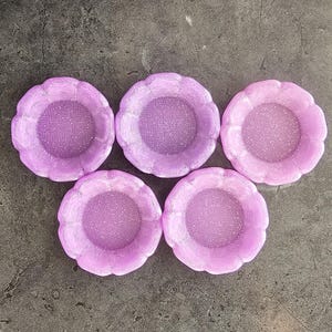 May include: Five lavender-colored, scalloped-edge dishes with a glittery interior are arranged on a dark gray surface. The dishes have a flower-like shape, with a textured, raised rim. The center of each dish has a sparkly finish.