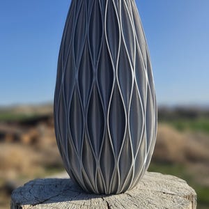 May include: A gray ceramic vase with a unique, textured, woven design. The vase is sitting on a weathered wooden stump.