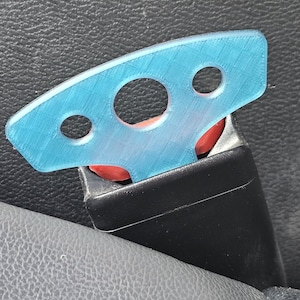 May include: A light blue plastic seat belt buckle guard with three holes. The guard is inserted into a black seat belt buckle, with a red release button visible. The background is a textured gray surface.