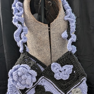 May include: A handmade crochet top in black and lavender, adorned with floral and butterfly motifs. The top features a unique design with a ruffled lavender trim and is displayed on a mannequin.