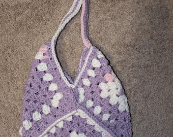 Puede incluir: Bolso tote de crochet hecho a mano con un patrón de cuadrados de la abuela. El bolso presenta una base de color lila claro con detalles blancos y rosas. La correa es una combinación de blanco, lila claro y rosa. El bolso tiene una abertura en forma de V.