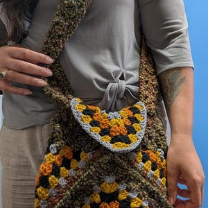 Może przedstawiać: Ręcznie robiona torba na ramię z szydełka z kolorowym wzorem granny square. Torba ma mieszankę pomarańczowych, żółtych, czarnych i szarych kwadratów z brązowym i zielonym paskiem. Torba jest noszona na ramieniu.