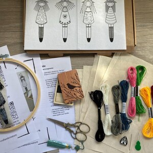 May include: An embroidery kit featuring a wooden hoop, printed patterns, embroidery floss in various colours, scissors, and a bookmark. The kit includes a box with four doll designs and instructions. The bookmark has a witch doll design.