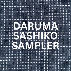 May include: A dark blue fabric featuring a repeating white cross-stitch pattern across the entire surface. The text DARUMA SASHIKO SAMPLER is printed in large, bold, white capital letters across the centre of the fabric.