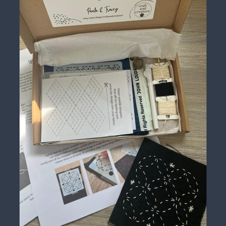May include: An open cardboard box containing a craft kit. Inside are spools of thread, a needle, a pen, and printed instructions. The kit includes a black fabric square with a white embroidered design and a white paper with a geometric pattern.