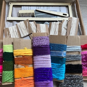 May include: A craft kit with a wooden loom, comb, and weaving tool. The kit includes spools of yarn in various colors, including pink, green, yellow, orange, purple, blue, gray, and white. The kit is packaged in a cardboard box.
