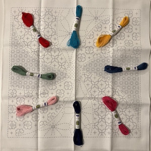 May include: A white fabric square featuring a printed grey geometric and floral pattern for embroidery, accompanied by seven individual skeins of embroidery floss in red, light blue, yellow, green, dark blue, pink, and dark magenta colours.