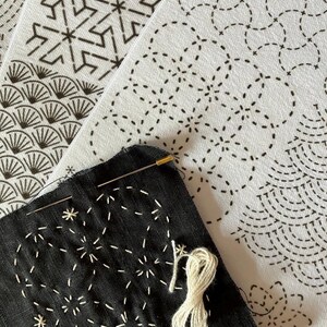 May include: Close-up of a black fabric with white starburst embroidery, a needle, and a spool of white thread. Several white fabric panels with black geometric patterns are in the background, showcasing a craft project.