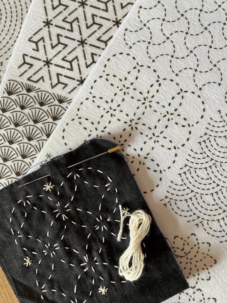 May include: Close-up of a black fabric with white starburst embroidery, a needle, and a spool of white thread. Several white fabric panels with black geometric patterns are in the background, showcasing a craft project.