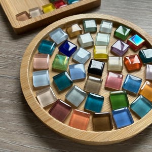 May include: A collection of colorful, square glass tiles in various shades of blue, green, pink, yellow, and brown. The tiles are arranged on a round, wooden tray and a rectangular wooden tray. The tiles are likely for a mosaic project.