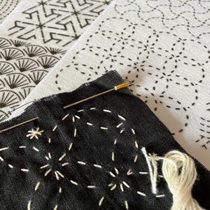 May include: Close-up of a black fabric with white stitching, showcasing a starburst pattern. Two needles are inserted, with a spool of white thread nearby. In the background, there are white fabrics with black geometric patterns, suggesting a craft project.