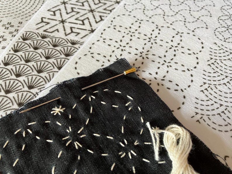 May include: Close-up of a black fabric with white stitching, showcasing a starburst pattern. Two needles are inserted, with a spool of white thread nearby. In the background, there are white fabrics with black geometric patterns, suggesting a craft project.