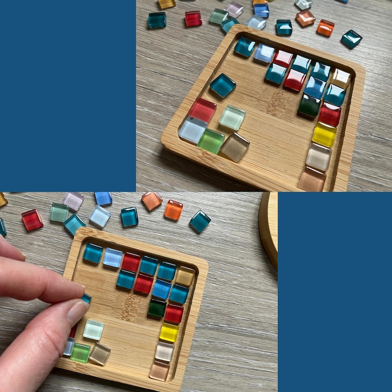 May include: A wooden tray with a grid pattern filled with colorful, square glass tiles. The tiles are in shades of red, blue, green, yellow, and orange. Some tiles are scattered around the tray. A hand is placing a blue tile into the tray.