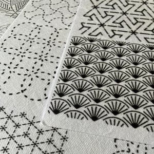 May include: Close-up of several white fabric pieces with black geometric patterns. The designs include fan shapes, dotted lines, and starbursts. The fabric has a textured appearance, and the patterns are neatly printed.