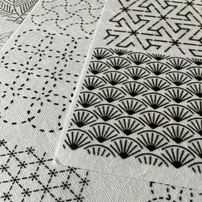 May include: Close-up of several white fabric pieces with black geometric patterns. The designs include fan shapes, dotted lines, and starbursts. The fabric has a textured appearance, and the patterns are neatly printed.
