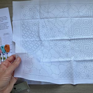 May include: A white fabric square featuring pre-printed grey geometric Sashiko embroidery patterns, accompanied by a set of six colourful embroidery floss skeins and an instructional paper titled Stitch Through Time with text details.