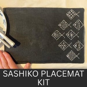 May include: Black rectangular placemat with white Sashiko fish pattern stitching. The placemat is on a white surface. The text "SASHIKO PLACEMAT KIT" is at the bottom. A plate and cutlery are visible.