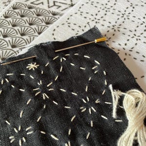 May include: Close-up of black fabric with white hand-stitched patterns, featuring a needle with a gold head. A bundle of white thread is visible, suggesting a craft project. The background shows patterned fabric.