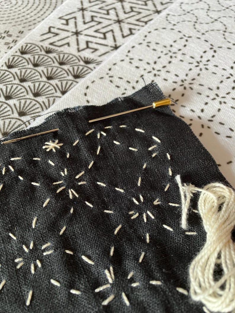 May include: Close-up of black fabric with white hand-stitched patterns, featuring a needle with a gold head. A bundle of white thread is visible, suggesting a craft project. The background shows patterned fabric.