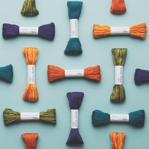May include: A collection of Sashiko thread skeins in various colours including purple, teal, orange, and olive green. Each skein is wrapped with a white paper band labelled Sashiko Thread 100m, arranged in a grid pattern on a light blue background.