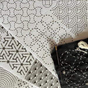 May include: Close-up of various white fabric pieces with black stitched patterns, including geometric and floral designs. A black fabric piece with white stitching, a needle, and white thread are also visible, suggesting a craft project.