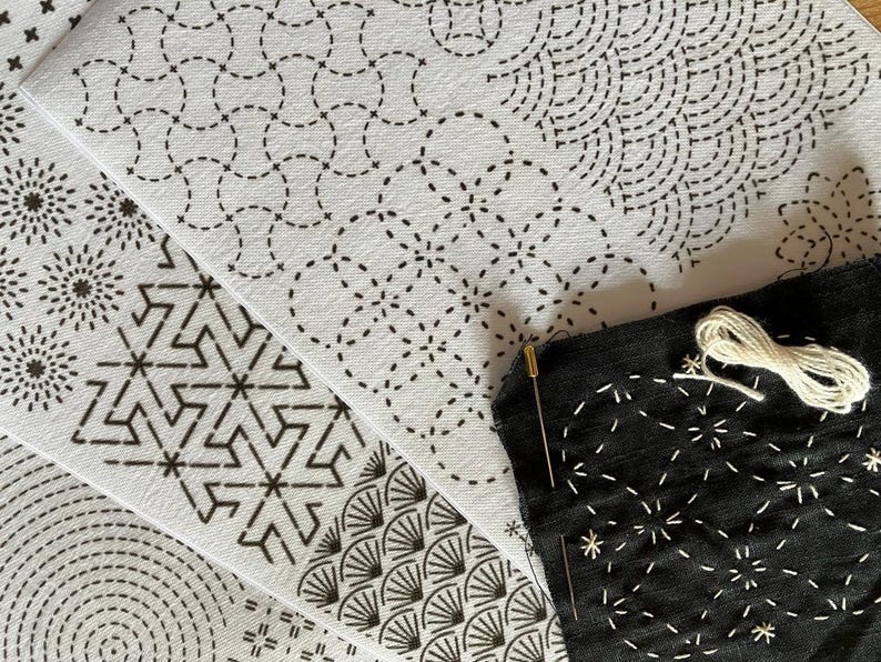 May include: Close-up of various white fabric pieces with black stitched patterns, including geometric and floral designs. A black fabric piece with white stitching, a needle, and white thread are also visible, suggesting a craft project.