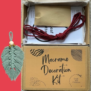 Peut inclure: Un kit de décoration macramé dans une boîte en carton, comprenant un ornement de feuille verte terminé avec une perle en bois et des accents dorés. Le kit comprend du cordon rouge et des instructions. La boîte est étiquetée "Macramé Decoration Kit".