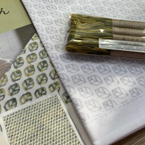 Könnte beinhalten: Eine Sammlung von Stoffen und Stickzubehör. Weißer Stoff mit einem grauen Blattmuster ist zu sehen, zusammen mit einer Packung grün-gelben Fadens mit der Aufschrift "Sashiko". Ein kleines, genähtes Stück und ein Buch sind ebenfalls vorhanden.