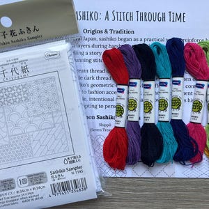 May include: A Sashiko sampler kit featuring a white fabric panel with printed geometric patterns, labelled Chiyogami, alongside eight skeins of embroidery thread in red, purple, dark blue, teal, navy, pink, light green, and yellow, plus an instruction sheet.