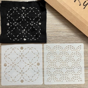 May include: A black fabric square with white stitched floral design, two white stencils with dotted patterns, and a brown cardboard box. The stencils feature geometric and wave-like designs. The kit is for crafting.