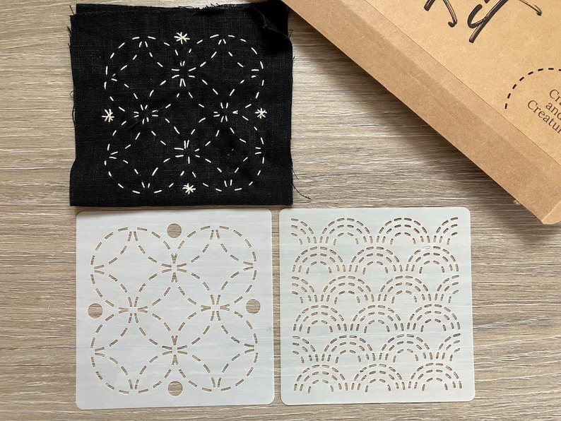 May include: A black fabric square with white stitched floral design, two white stencils with dotted patterns, and a brown cardboard box. The stencils feature geometric and wave-like designs. The kit is for crafting.